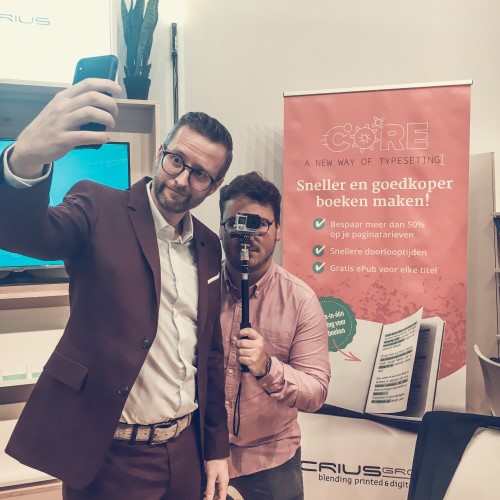 Twee mannen maken een selfie bij een Crius Group-banner op een uitgeversbijeenkomst.