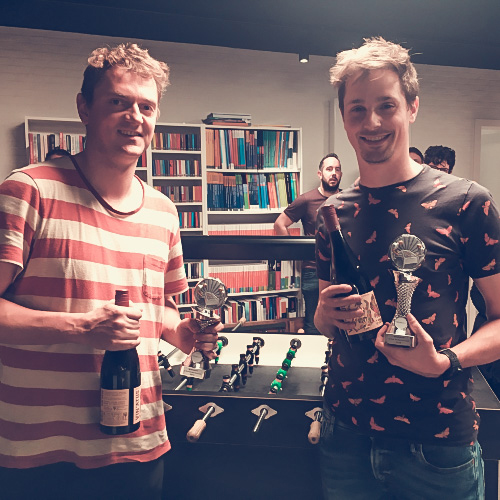 Twee personen poseren met bekers en een fles voor een tafelvoetbaltafel in een kamer met boekenplanken.