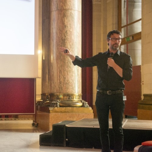 Spreker met microfoon presenteert voor een publiek in een grote zaal met pilaren en een scherm.