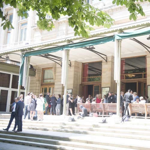 Mensen staan op de trappen voor een gebouw met grote zuilen onder een zonnige hemel.