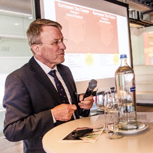 Man geeft presentatie bij ronde tafel met microfoon en projectiescherm op de achtergrond.
