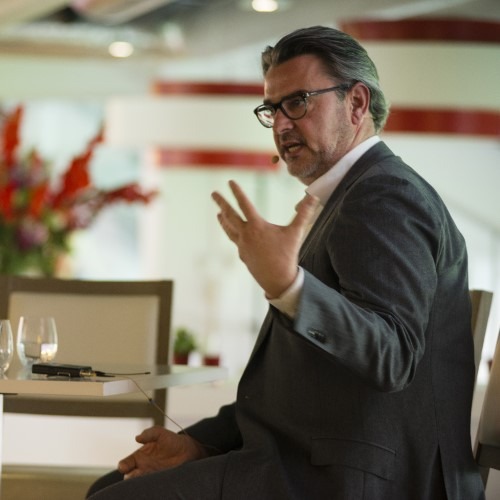 Man in pak geeft presentatie en maakt handgebaren, met bloemen op de achtergrond.