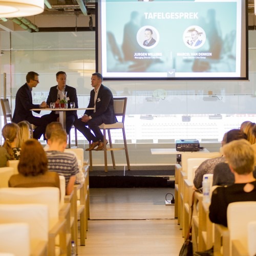Paneldiscussie op podium met publiek, projectiescherm toont presentatie in conferentieomgeving.