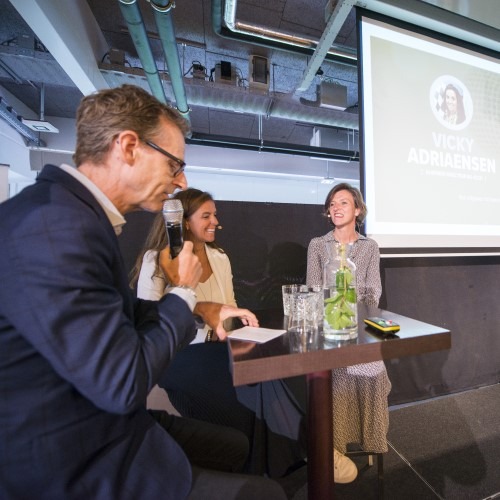 Drie personen in gesprek tijdens een presentatie op een podium met microfoon en projectiescherm.