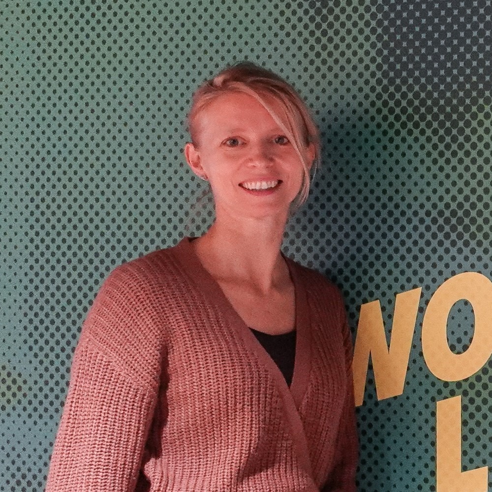 Woman in brown cardigan smiles in front of green backdrop with yellow letters.