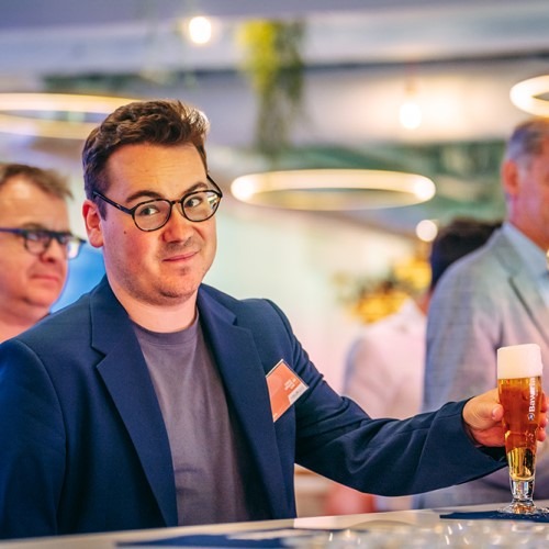 Man in blauw colbert met bril houdt een glas bier vast tijdens een evenement.