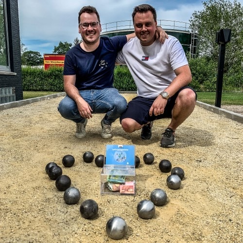 Twee mannen poseren naast een hartvorm van petanqueballen op een veld.