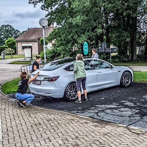 Mensen duwen een elektrische auto op een parkeerplaats bij een laadstation in een groene buurt.