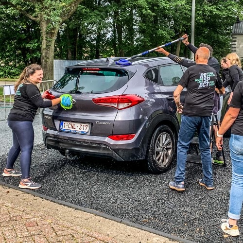 Groep mensen wast gezamenlijk een auto op een buitenlocatie omgeven door bomen.