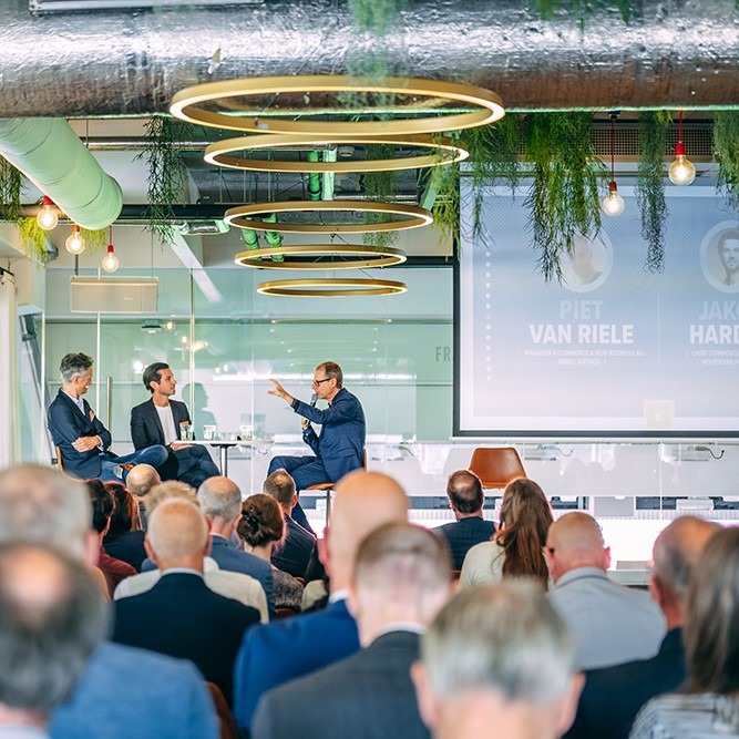 Zakelijke presentatie met publiek, spreker wijst naar scherm in moderne, groene ruimte.