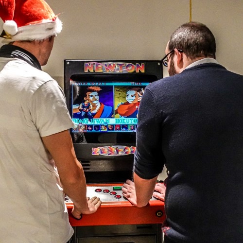 Twee mannen spelen op een retro-arcadekast, eentje met een kerstmuts. Gezelligheid tijdens een spelmiddag.