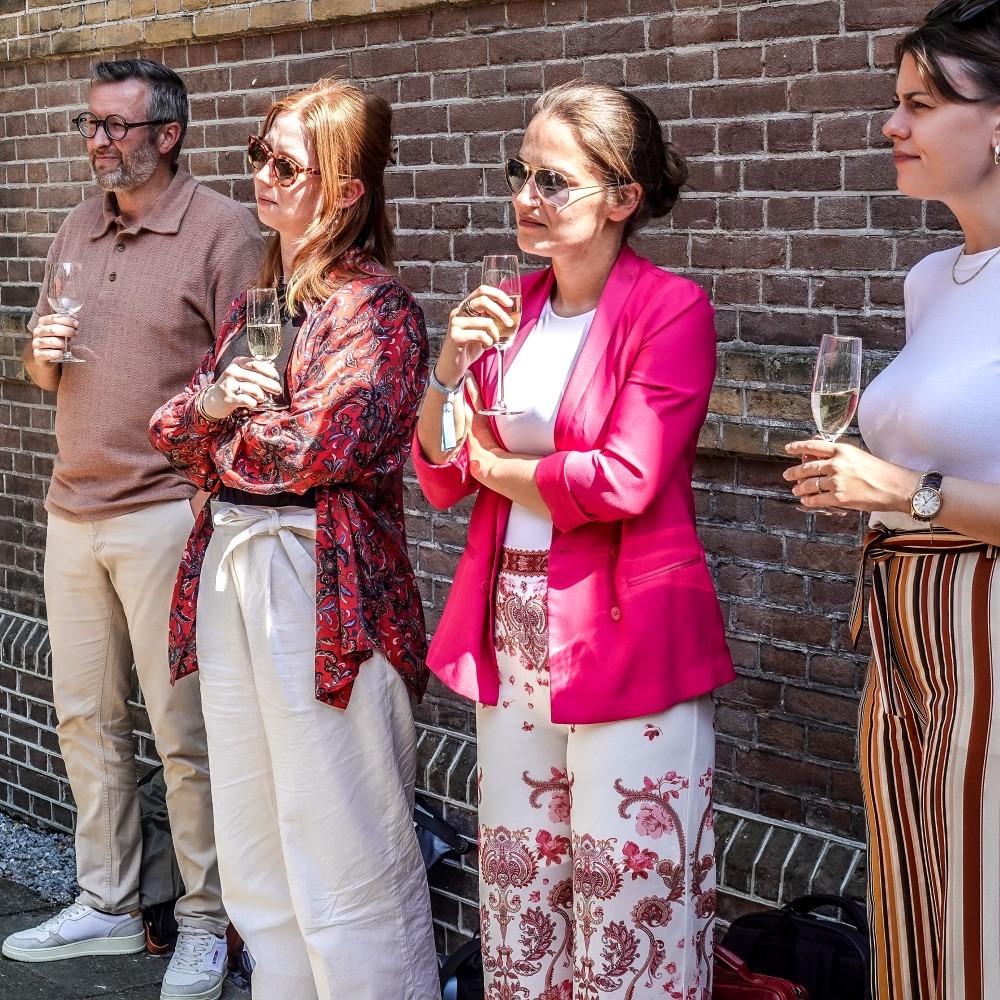 Group of people outside in casual clothing with drinks, smiling and talking in front of a brick wall.