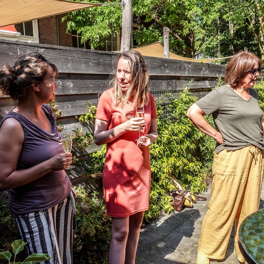 Three people stand outside talking and drinking in a garden next to a wooden fence.