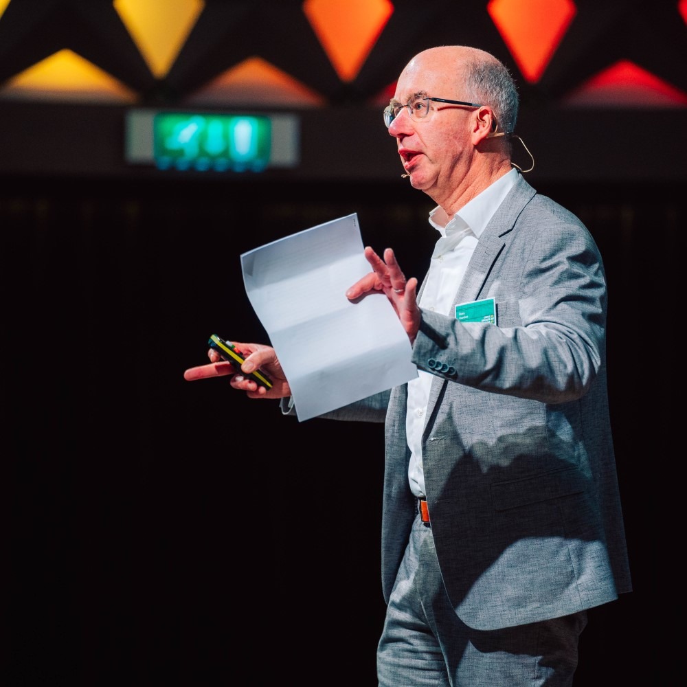 Man in pak spreekt op podium met papieren in de hand, kleurrijke achtergrond.