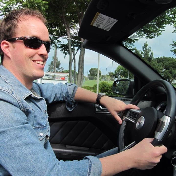 Man wearing sunglasses drives a convertible car on a sunny day.