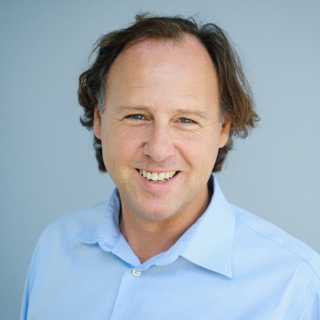 Man met donkerblond haar, lachend en een lichtblauwe blouse dragend, voor een effen achtergrond.