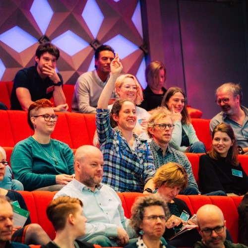 A group of people seated in an auditorium with one person raising their hand during a presentation.