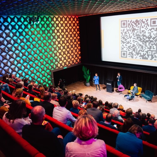 A presentation in an auditorium with a large QR code on screen and colorful lighting on the side walls.