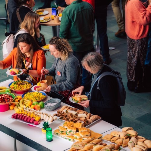 People gathering food at a buffet during an event, featuring colorful dishes and fruit.