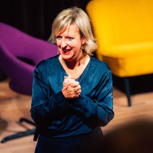Woman in a blue blouse speaking on stage with purple and yellow chairs in the background.